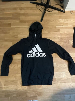 Svart hoodie från Adidas med logga - Svart hoodie från Adidas med stor vit logga på bröstet. Klassisk huva med snörning och ribbade muddar vid ärmar och nederkant. Perfekt för en avslappnad och sportig stil.