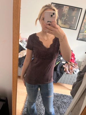 Mörkbrun spetsdetaljerad topp - Söt mörkbrun topp med v-ringning och spetsdetaljer längs halsringningen. Kortärmad och figurnära passform, perfekt att styla med jeans eller kjol. Materialet känns mjukt och lätt mot huden.