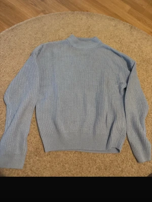 Ljusblå ribbstickad tröja - En stilren ljusblå stickad tröja med ribbad struktur och högre hals. Tröjan har långa ärmar och breda muddar vid ärmslut och nederkant. Perfekt för lager på lager och enkel att matcha med olika outfits.