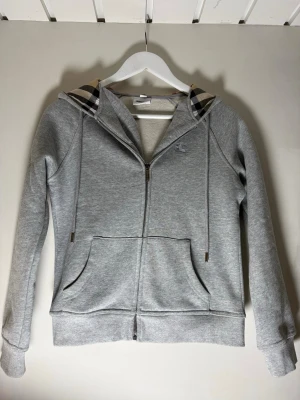 Grå zip hoodie från Burberry - Snygg grå zip hoodie från Burberry med klassiskt rutigt foder i huvan och dragsko. Hoodien har två stora fickor fram och ribbade muddar vid ärmar och nederkant. Perfekt för en chill och stilren look. Tillverkad i mjuk bomullsblandning.