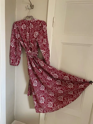 Rödvit klänning stl. XS - Snygg röd klänning med vita blommiga och spiralformade mönster. Klänningen har lång bred ärm, knyt i midjan och en svepande kjol som ger en feminin siluett. Svagt puffärm enligt bild. Perfekt till somriga grillfester. Stl. XS från Pigalle. Använd 1 gång ett tidigt firande. Hel o ren. Som ny. Passar dock inte min skära hudton. Tunn men inte så man ser igenom tyget. 