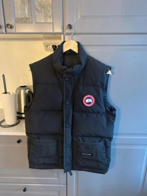 Svart dunväst från Canada Goose - Snygg svart dunväst från Canada Goose med hög krage och två stora fickor framtill. Västen har det klassiska runda Canada Goose-märket på bröstet och stängs med både dragkedja och knappar. Perfekt för lager-på-lager och håller dig varm under kyliga dagar.