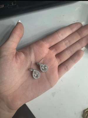Silvriga droppformade örhängen - Säljer ett par silvriga örhängen med droppformad design. Örhängena har en stor klar sten i mitten omgiven av mindre gnistrande stenar, vilket ger en riktigt lyxig känsla. Perfekta för dig som gillar bling och vill sticka ut lite extra.