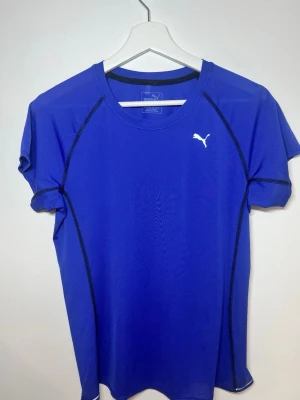 Blå träningströja från Puma - Sportig blå t-shirt från Puma med svarta sömmar och vit logga på bröstet. Tillverkad i ett lätt och ventilerande material som passar perfekt för träning och aktivitet. Klassisk rund halsringning och korta ärmar.