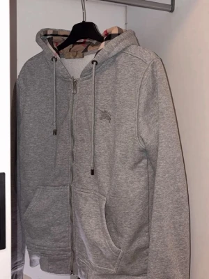 Grå zip hoodie från Burberry - Snygg grå zip hoodie från Burberry med klassiskt rutigt foder i huvan och broderad logga på bröstet. Hoodien har dragsko i huvan, två stora fickor fram och är gjord i mjukt bomullsmaterial. Perfekt för en chill och stilren look.