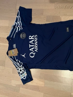 PSG matchtröja Barcola #29 blå - Snygg mörkblå PSG matchtröja från Jordan med Barcola och nummer 29 på ryggen. Tröjan har vita detaljer, Champions League-märke på ärmen och Qatar Airways som sponsortryck. Tillverkad i lätt och ventilerande material, perfekt för fotboll eller träning. Storlek S 