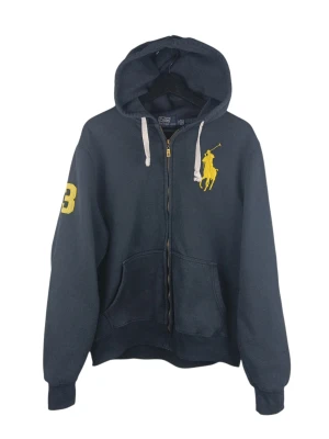 Vintage Polo Ralph Lauren Zipper marinblå huvtröja, XL - Marinblå huvtröja från Polo by Ralph Lauren med dragkedja. Tröjan har en stor gul broderad logotyp på bröstet som föreställer en polospelare till häst. På höger ärm finns ett stort gult nummer '3' broderat. Huvtröjan har en huva med dragsko och två fickor framtill. Inga hål eller slitage. Armhåla till armhåla: 59cm Axelbredd: 48cm Långt: 66cm Armlängden från armhåla: 54cm