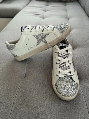 Golden Goose sneakers med glitter - Snygga vita Golden Goose sneakers med silverglittriga detaljer på tå, häl och stjärna på sidan. Skorna har vita snören med svart text och en klassisk låg siluett. Tillverkade i läder med robust gummisula. Perfekta för dig som vill sticka ut.