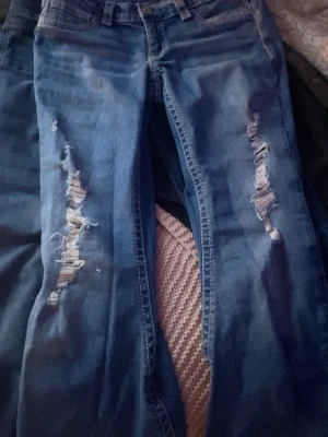 Blå slitna jeans med hål - Säljer ett par blå jeans med slitningar och hål på båda benen. Jeansen har klassisk femficksmodell och en snygg detalj på bakfickan med dekorativ söm och knapp. Materialet är denim i bomull och passformen är smal.