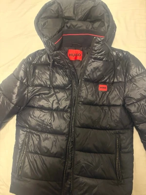 Svart pufferjacka från Hugo Boss - Svart pufferjacka från Hugo Boss med röd logotyp på bröstet och insidan. Jackan har en stor huva, dragkedja framtill och två sidofickor med dragkedja. Materialet är glansigt och quiltat, vilket ger en modern look och extra värme under vintern. Jackan funkar även utmärkt under vår och höst!