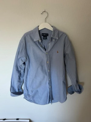 Blå skjorta från Ralph Lauren - Säljer en klassisk ljusblå skjorta från Ralph Lauren med broderad logga på bröstet. Skjortan har knappar framtill, långärmad design och är tillverkad i bomull. Perfekt för dig som gillar stilrena plagg med ikonisk branding.
