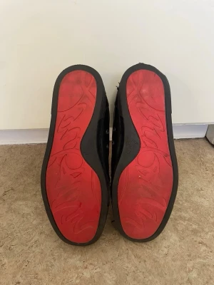 Svarta sneakers med nitar från Louboutin - Säljer ett par svarta sneakers från Christian Louboutin med röda sulor och coola nitar på tån. Skorna har en låg profil och är tillverkade i läder med metalliska detaljer. Perfekt för dig som vill sticka ut med en edgy och lyxig stil.