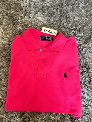 Rosa polotopp från Polo Ralph Lauren - Säljer en klassisk rosa polotopp med ikonisk broderad logga på bröstet. Toppens material är bomull och den har krage samt knappar framtill. Perfekt för dig som gillar stilrena och färgstarka plagg.
