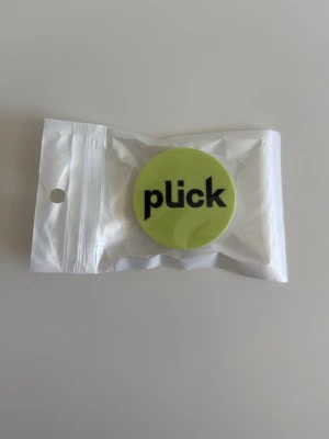 Plick-popsocket - Säljer en rund popsocket med ljusgrön/gul bakgrund och svart text där det står 'plick'. Helt oanvänd, kommer i en liten plastpåse.