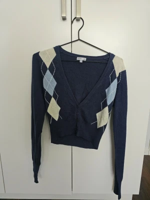 Mörkblå argyle kofta från Ellos - Mörkblå kofta från Ellos med argyle-mönster i ljusblått och beige på axlarna. Djup v-ringning, knäppning framtill och långa ärmar. Perfekt för att skapa en preppy look.