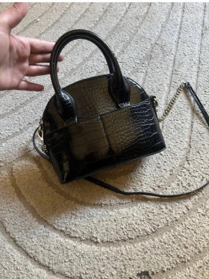 Svart croco-mönstrad handväska - Säljer åt en kompis😊Cool svart handväska med croco-mönster och glansig finish. Väskan har två korta handtag och en lång axelrem med kedjedetaljer. Perfekt för dig som gillar trendiga accessoarer och vill sticka ut med en unik look. Finns ej att köpa längre därav priset💕