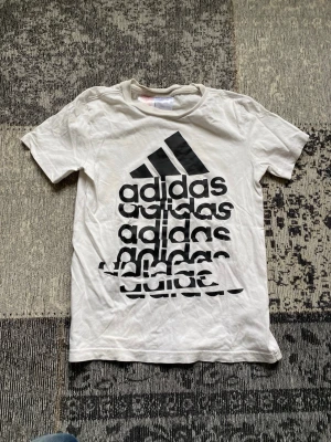 Vit Adidas t-shirt med tryck - Säljer en vit t-shirt från Adidas med stort svart logotryck på framsidan där Adidas-loggan och texten upprepas i en cool design. T-shirten har korta ärmar och klassisk passform. Perfekt för dig som gillar sportiga och stilrena plagg.