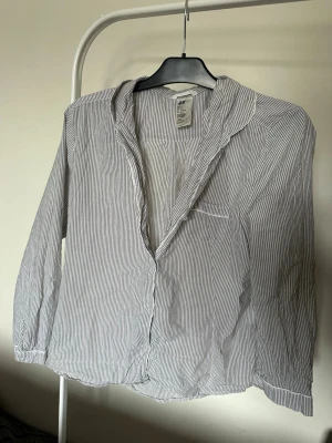 Randig pyjamasskjorta från H&M - Klassisk pyjamasskjorta från H&M i vitt med tunna grå ränder. Skjortan har lång ärm, krage och bröstficka. Tillverkad i mjukt och lätt material, perfekt för avslappnade kvällar hemma.