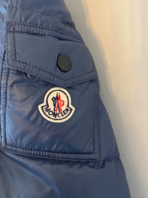 Blå dunjacka från Moncler - Snygg blå dunjacka från Moncler med klassisk logotyp på ärmen och praktisk ficka med knapp. Jackan har quiltad design och dragkedja framtill. Perfekt för kalla vinterdagar och tillverkad i glansigt material som ger en exklusiv look.