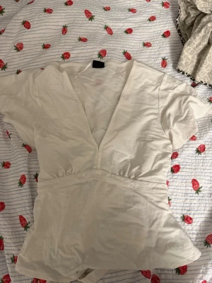 Vit omlottopp från Gina Tricot - Säljer en vit omlottopp från Gina Tricot i storlek S. Toppen har korta ärmar, djup v-ringning och markerad midja som ger en snygg siluett. Perfekt att matcha med jeans eller kjol för en fräsch look.