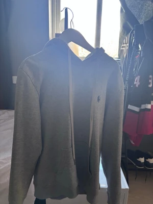 Grå hoodie från Polo Ralph Lauren - Säljer en klassisk grå hoodie från Polo Ralph Lauren med broderad logga på bröstet. Hoodien har huva med dragsko, magficka och ribbade muddar. Perfekt för en avslappnad och stilren look. Tillverkad i mjuk bomullsblandning.