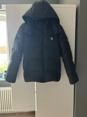 Svart pufferjacka från Calvin Klein - Svart pufferjacka från Calvin Klein med huva och dragkedja. Jackan har CK logga på bröstet och en patch på ärmen. Fylld för extra värme och har två fickor med dragkedja.