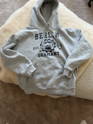 Grå hoodie med Berlin-tryck - Säljer en grå hoodie med stor huva och känguruficka. Tryck på bröstet med texten 'Berlin Germany' och emblem. Perfekt för dig kring 170-180 str S-XS