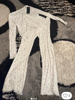 Vit blommig pyjamas med lång ärm - Supermysig vit pyjamas från New yorker med små blå blommor. Setet har långärmad topp med knappar och matchande byxor med utsvängda ben. Materialet är mjukt och stretchigt, perfekt för chillkvällar hemma.