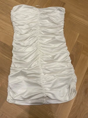 Vit axelbandslös kortklänning Gina Tricot - Säljer en vit axelbandslös kortklänning från Gina Tricot. Klänningen har snygg rynkad design framtill och är gjord i ett glansigt material som ger en lyxig känsla. Perfekt för dig som vill sticka ut med en stilren look.