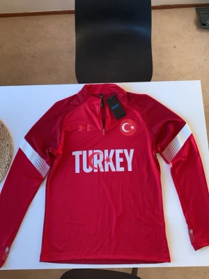 M. Turkisk träningströja  - Köpte den för 1 månad sen med prislapp från en svensk företag game changer Jersey hade ingen aning om att deras tillverknings fabrik va i Kina så när jag köpte XL kom hem som M visste inte att kinesisk storlek är 2 storlekar mindre i Europa röd långärmad träningsjacka tryck Under Armour med halv dragkedja och tryck 'TURKEY' på bröstet. Träningströjan har vita detaljer på ärmarna och Turkiets flagga tryckt på vänster sida. Materialet är syntetiskt och passar perfekt för sportaktiviteter.