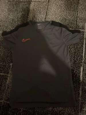 Mörkgrå Nike t-shirt med svart detalj - Snygg mörkgrå t-shirt från Nike med svarta detaljer på axlarna och röd Nike-logga på bröstet. Tillverkad i ett mjukt och lätt material som passar perfekt till träning eller chill. Klassisk passform och korta ärmar.