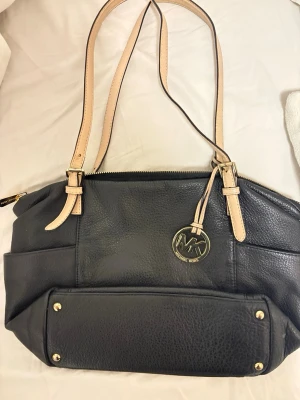Svart Michael Kors handväska i skinn - Snygg svart handväska från Michael Kors i äkta skinn med beige axelband och guldfärgade metalldetaljer. Väskan har ett klassiskt MK-hänge, dragkedja upptill och flera fack på utsidan. Perfekt för dig som vill ha en stilren och rymlig väska med exklusiv känsla.