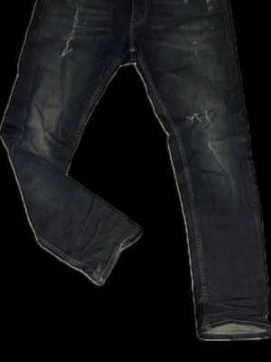  slitna replay jeans - Säljer ett par mörkblå jeans med slitningar och raka ben. Jeansen har snygga detaljer med slitna partier framtill och bakfickor med dekorativa sömmar. Perfekta för en avslappnad och trendig stil.