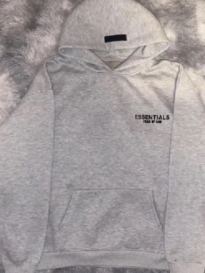 Grå Essentials Fear of God hoodie - Snygg grå hoodie från Essentials Fear of God med svart logotyp på bröstet och ryggen. Hoodien har en stor huva och klassisk känguruficka framtill. Materialet är mjukt och bekvämt, perfekt för en avslappnad streetwear-look.