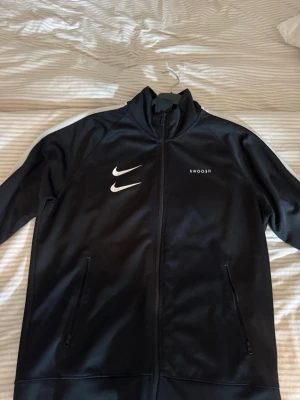 Svart Nike zip-jacka med Swoosh - Svart sportjacka från Nike med vit Swoosh-logga på både fram- och baksidan. Jackan har hel dragkedja, två fickor med dragkedja och texten 'SWOOSH' på bröstet. Materialet är slätt och syntetiskt, perfekt för träning eller chill.