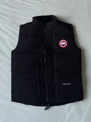 Svart dunväst från Canada Goose - Svart dunväst från Canada Goose med klassisk logotyp på bröstet och två stora fickor framtill. Västen har quiltad design, hög krage och dragkedja med tryckknappar. Perfekt för kalla dagar och snygg att matcha med hoodie eller t-shirt.