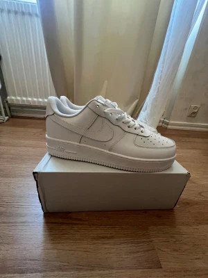 Nike Air Force 1 vita sneakers - Klassiska helvita Nike Air Force 1 sneakers i lågt utförande. Skorna har en stilren design med perforerad tå, Nike swoosh på sidorna och AIR-detalj på sulan. Tillverkade i läder med vit gummisula och vita skosnören. Perfekta för en clean och tidlös look.