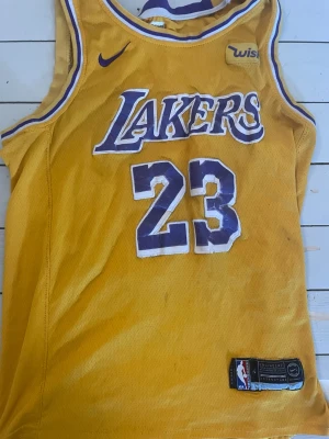 Gul Lakers basketlinne nr 23 - Säljer en gul basketlinne från Nike med Lakers-logga och nummer 23 på framsidan. På baksidan står det 'JAMES' och nummer 23. Linnet har lila och vita detaljer runt hals och ärmar, och är tillverkat i mesh-material som andas.