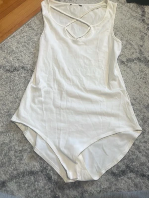 Vit ärmlös body med criss-cross - Snygg vit body i mjukt stretchigt tyg med ärmlös design och criss-cross detalj vid halsringningen. Perfekt att styla till jeans eller kjol för en clean look. Tryckknappar i grenen för enkel påklädning.