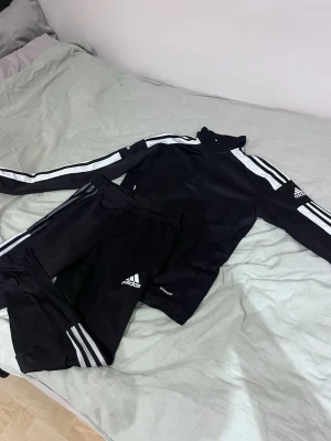 Svart Adidas träningsset med vita ränder - Snyggt svart träningsset från Adidas med klassiska vita ränder längs ärmar och ben. Setet består av en långärmad tröja med dragkedja i halsen och matchande byxor. Perfekt för träning eller chill. Tillverkad i ett mjukt och stretchigt material.