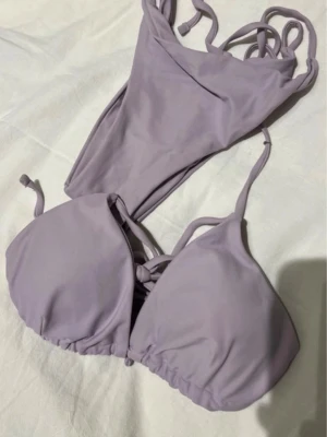 Ljuslila bikini med knyt - Säljer en snygg ljuslila bikini med knyt i både topp och trosa. Bikinin har en enkel och stilren design med triangelformade kupor och smala axelband. Perfekt för sommarens strandhäng eller poolparty!