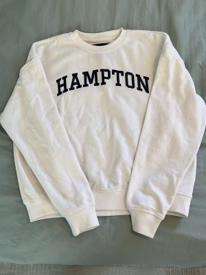 Vit sweatshirt med HAMPTON-tryck - Vit sweatshirt med svart HAMPTON-tryck över bröstet. Klassisk rund halsringning och ribbade muddar vid ärmslut och nederkant. Perfekt för en avslappnad och cool look. Materialet är mjukt och bekvämt, troligen bomull eller bomullsblandning.