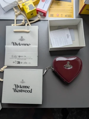 Hjärtfomad plånbok Vivienne Westwood - Säljer en hjärtformad plånbok från Vivienne Westwood i blankt vinrött läder med silverfärgad logga på framsidan. Plånboken har dragkedja och nyckelringar på insidan, perfekt för att hålla koll på nycklar och småsaker. Medföljer originalförpackning och påse.