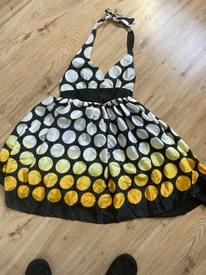Prickig halterneckklänning i svart och gult - Snygg kort halterneckklänning med stora vita och gula prickar på svart botten. Klänningen har knyt i nacken och markerad midja som ger en retroinspirerad look. Materialet känns glansigt och liknar polyester eller satin. Perfekt för dig som vill sticka ut!