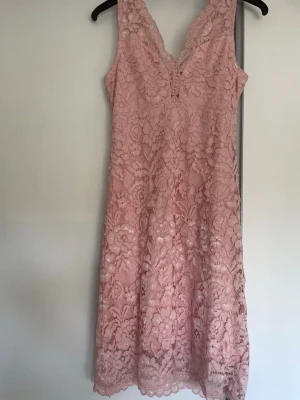 Rosa spetsklänning med dragkedja - Söt rosa klänning i spets med v-ringning och ärmlös design. Klänningen har ett blommigt spetsmönster och en synlig dragkedja i ryggen. Perfekt för sommarens alla tillfällen och ger en romantisk look.