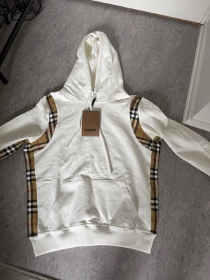 Vit Burberry hoodie - Säljer en vit hoodie från Burberry med ikoniska rutiga paneler längs sidorna och ärmarna. Hoodien har en stor huva med dragsko, en rymlig magficka och ribbade muddar. Materialet är mjukt och bekvämt, perfekt för en cool och avslappnad look.