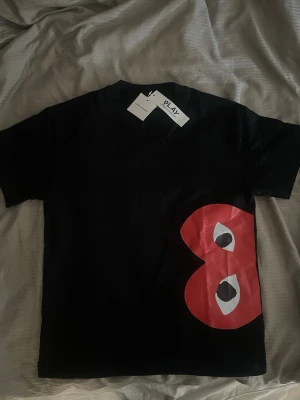Svart t-shirt från Comme des Garçons Play - Cool svart t-shirt från Comme des Garçons Play med stort rött hjärta och ögon på framsidan. Klassisk passform och korta ärmar, perfekt för dig som gillar streetstyle och grafiska prints.