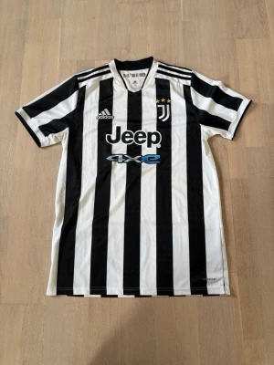 Juventus matchtröja Adidas M - Juventus officiella matchtröja från Adidas med klassiska svartvita ränder, logga och sponsortryck. Tröjan har tre stjärnor ovanför klubbmärket, rund hals och är tillverkad i Primegreen-material. Perfekt för fotboll och sport.