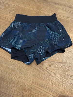 Svarta träningsshorts Nike XS - Säljer ett par svarta träningsshorts från Nike i storlek XS. Shortsen har ett blått och grönt geometriskt mönster och bred resår i midjan för extra komfort. Perfekta för gymmet eller löprundan.