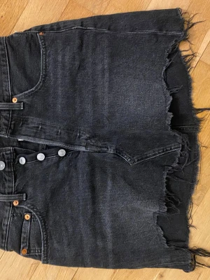 Svart jeanskjol från Levi's - Cool svart jeanskjol från Levi's med rå och fransig nederkant. Kjolen har klassiska fem fickor, silverfärgade knappar framtill och en snygg vintage-look. Perfekt för dig som gillar en edgy och avslappnad stil.